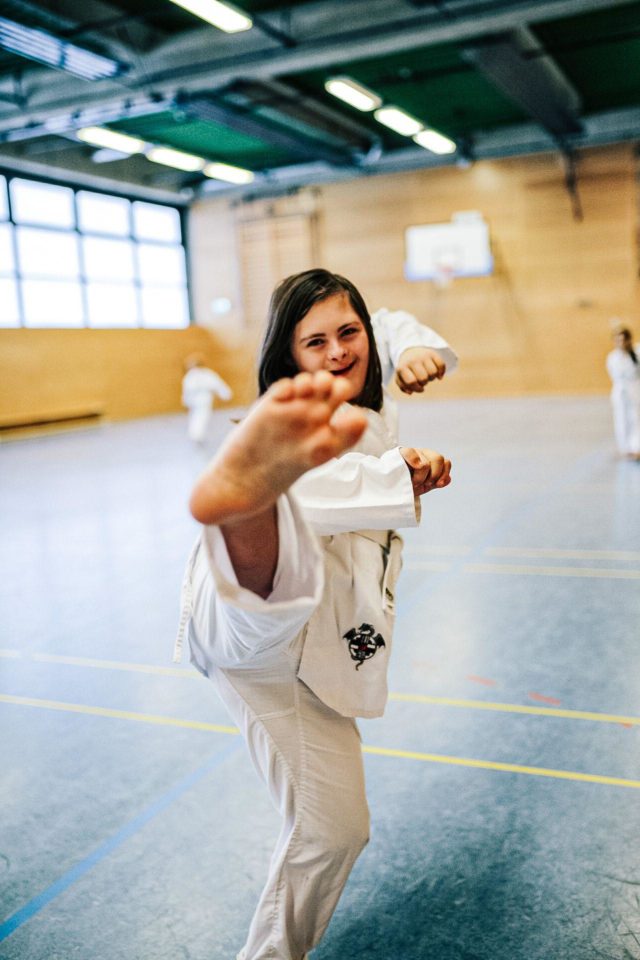 Mädchen mit Dow-Syndrom im Dobog (Taekwondo-Anzug). Sie tritt mit dem rechten Bein frontal nach oben Richtung Kamera. 