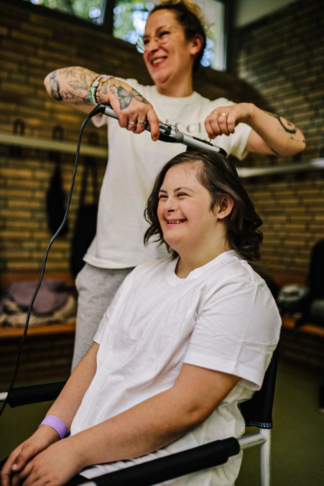 Teenangerin mit Down-Syndrom bekommt die Haare frisiert. Sie lacht gemeinsam mit der Stylistin in den Spiegel. Beide tragen ein weißes T-Shirt. 