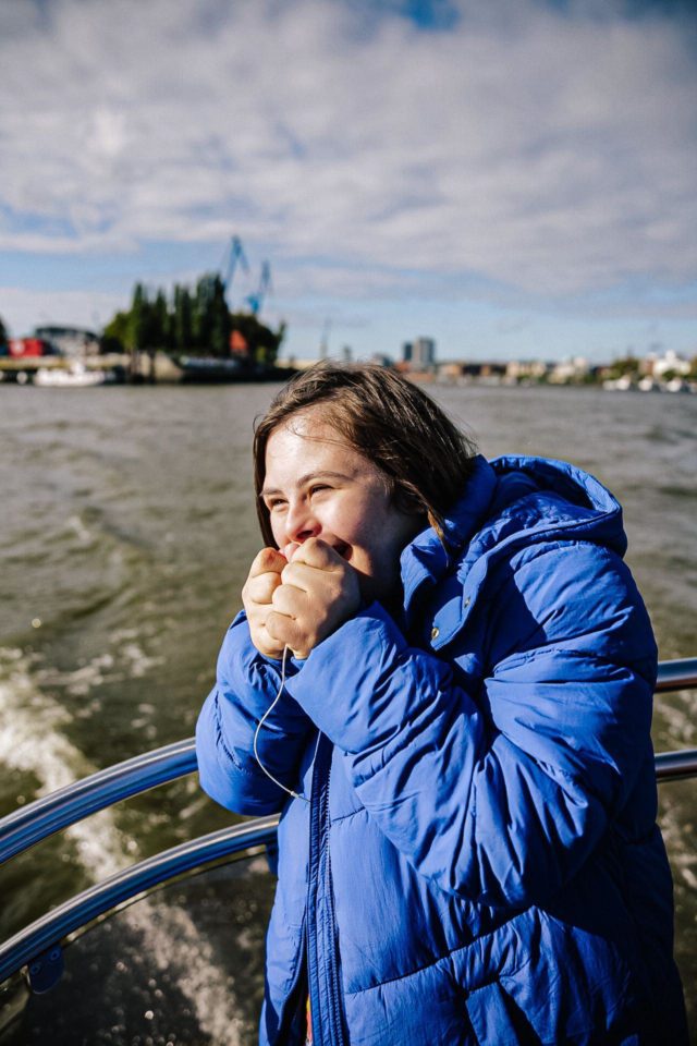Mädchen mit Donw-Syndrom und blauem Mantel steht an der Relining eines Schiffes. Sie hält die Hände in Fäusten lachend vor ihrem Gesicht. 